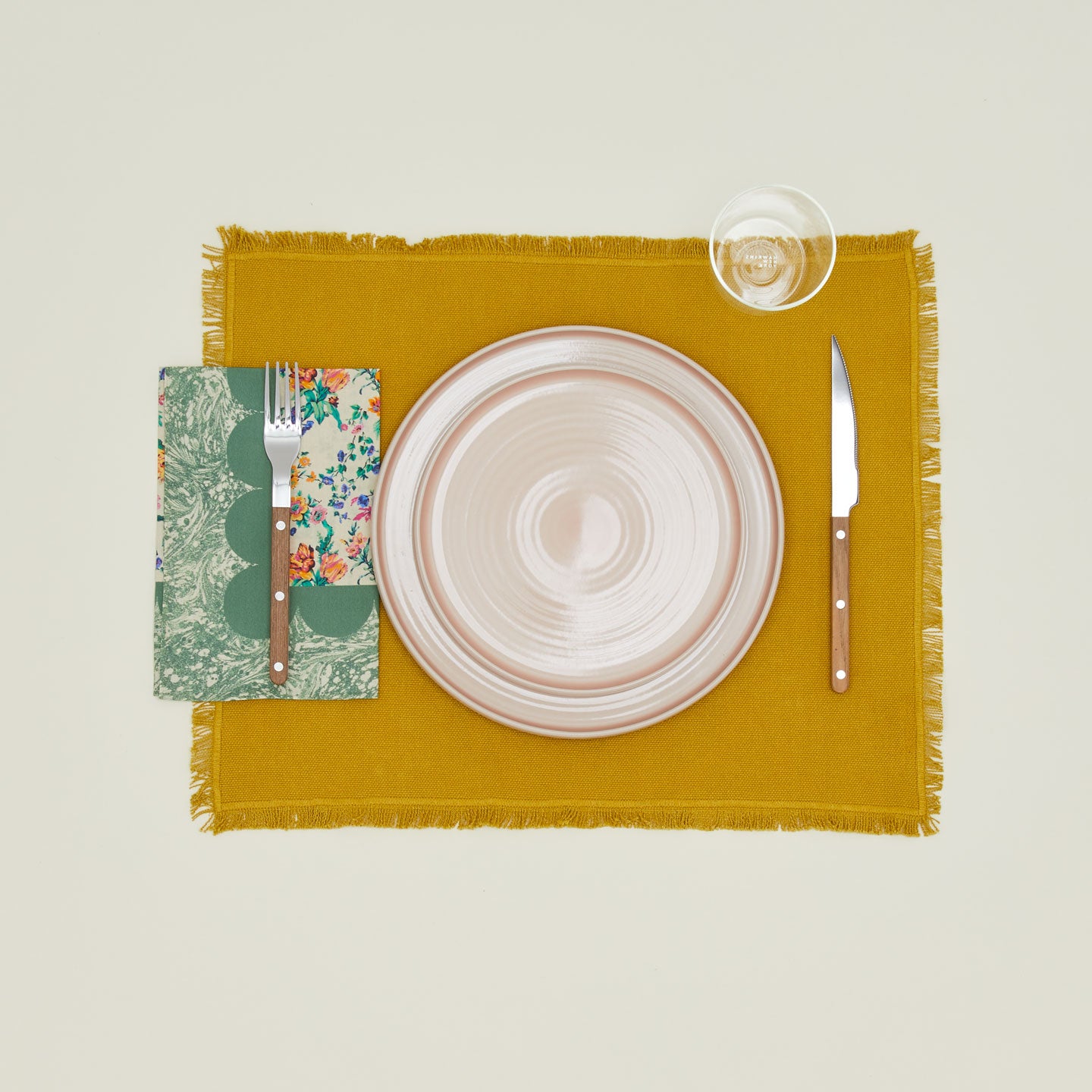 A table setting with blush plates, a clear glass, a mustard placemat, and a fern patterned napkin.