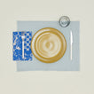 A table setting with mustard plates, a grey glass, a sky placemat, and a klein blue patterned napkin.