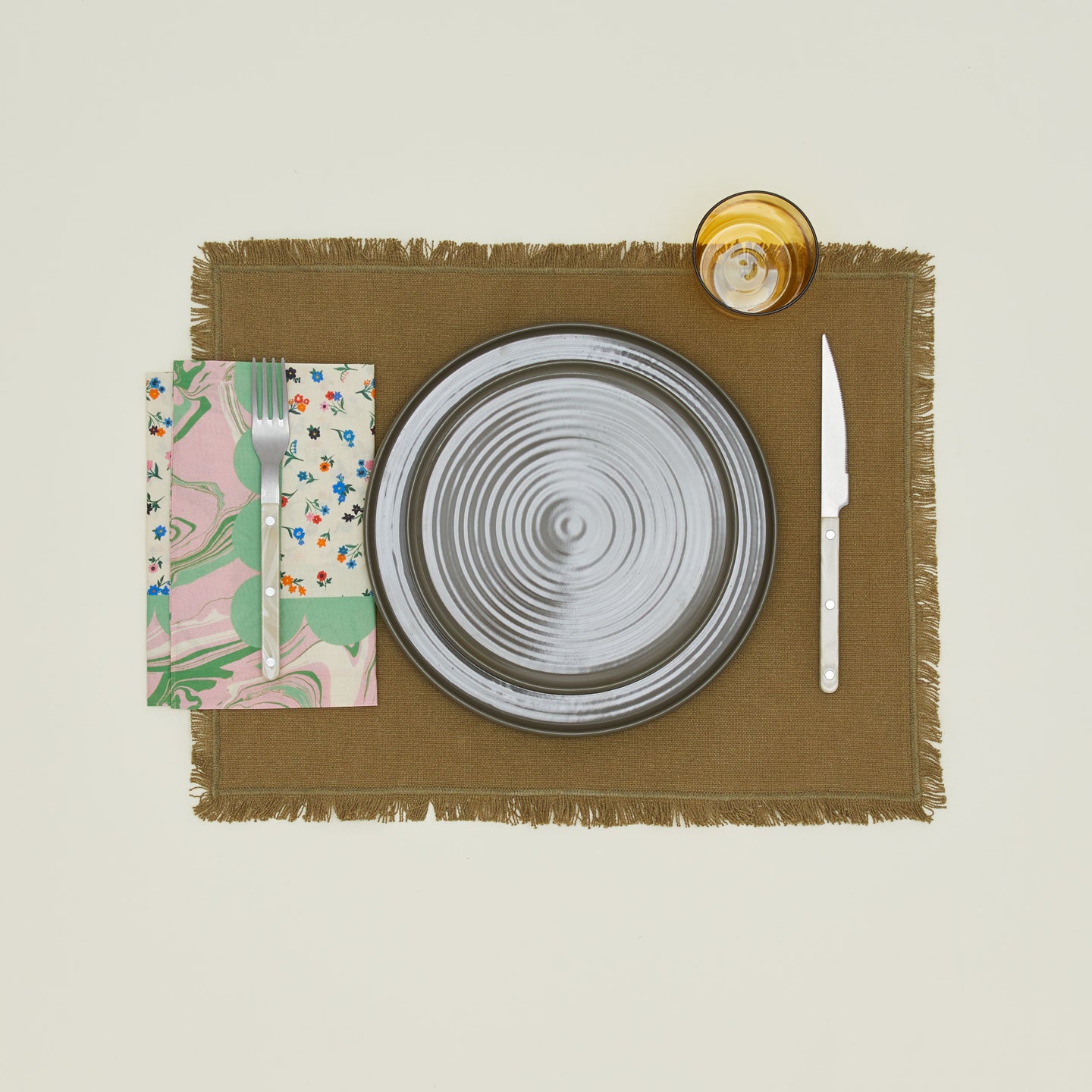 A table setting with olive plates, an amber glass, an olive placemat, and a fern patterned napkin.