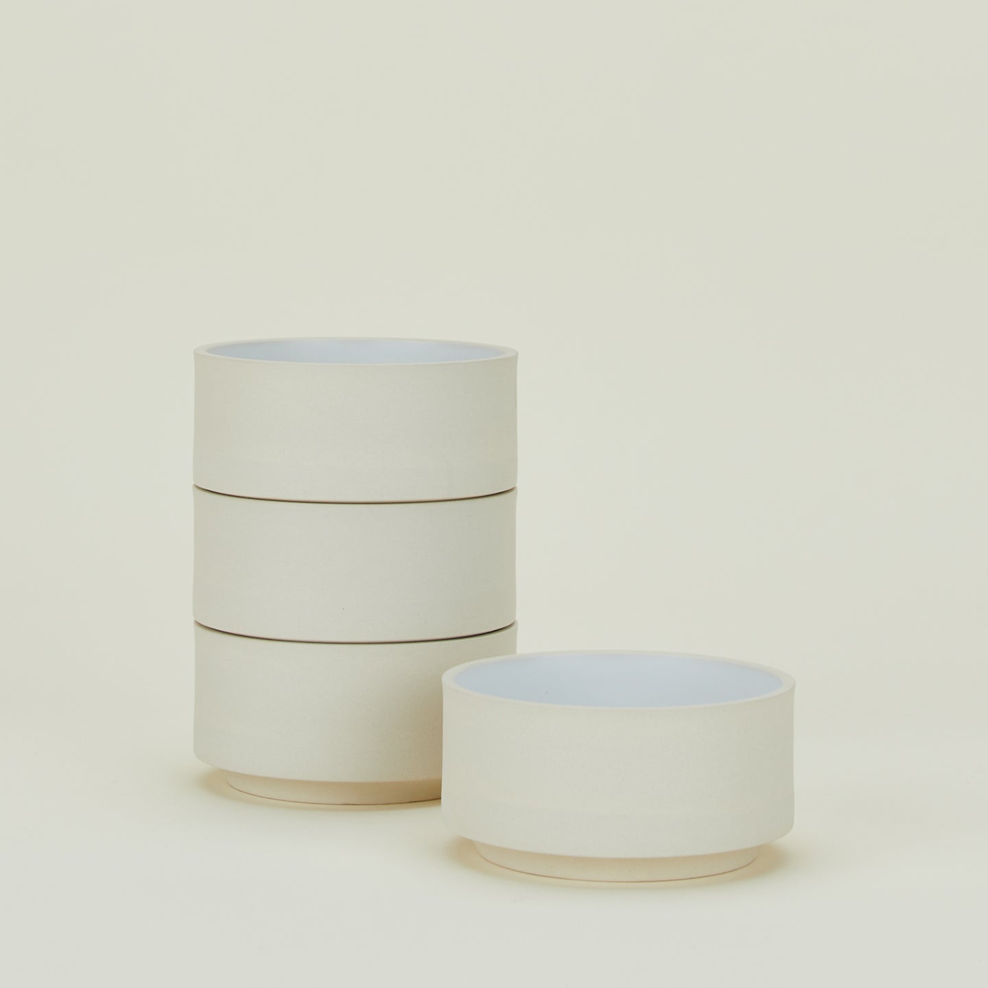 Four beige ceramic bowls are stacked vertically on the left, with a single bowl placed to the right on a light cream background.
