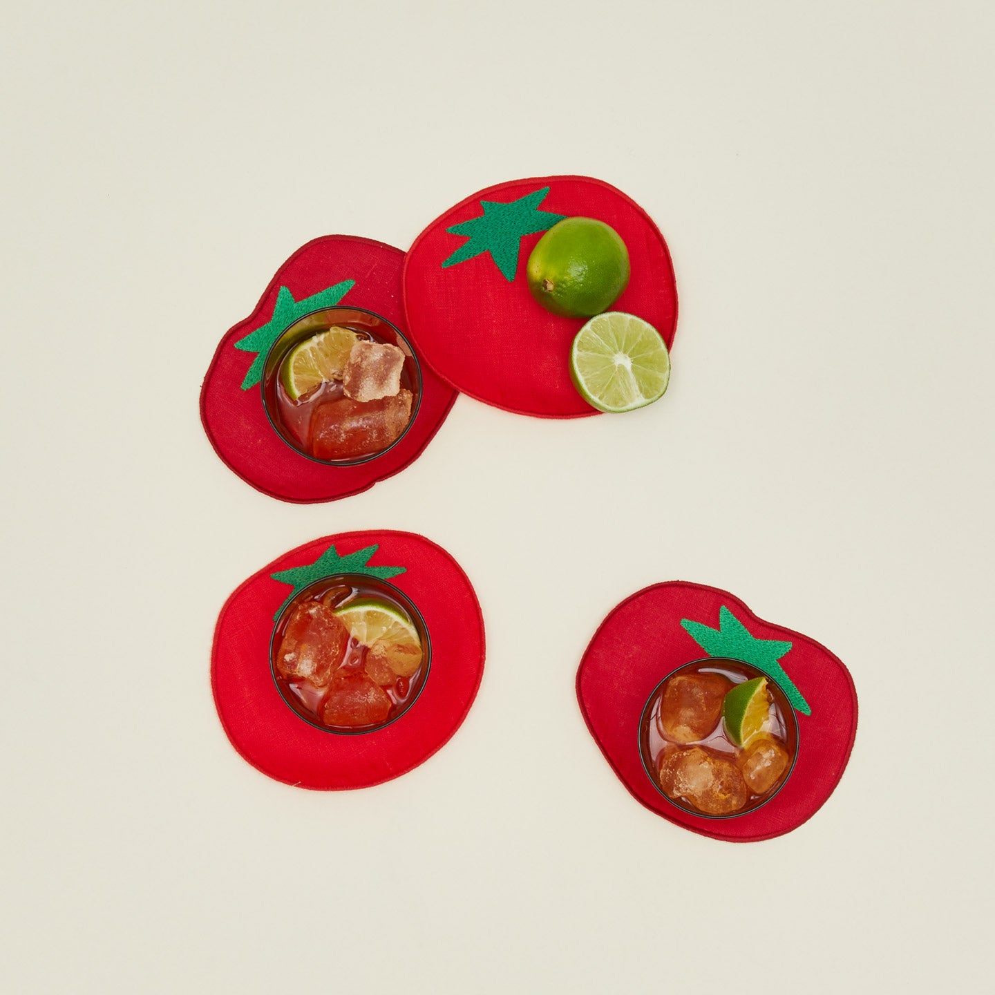 Four red tomato cocktail napkins with drinks and lime on a beige background