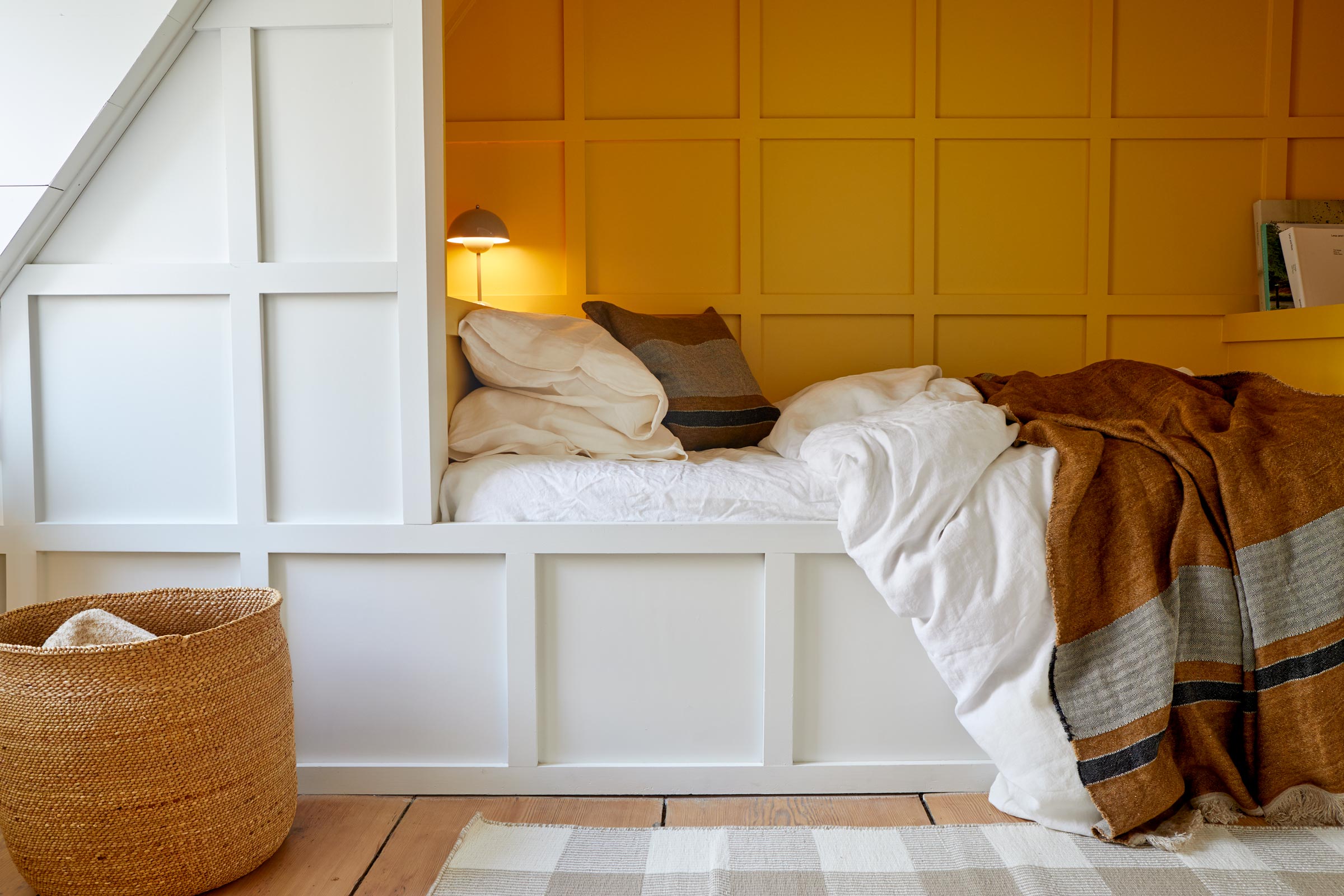 A cozy bed with white linens and pillows, complemented by a brown blanket is set against a paneled yellow wall. A lamp, basket, and white-painted wooden framing complete the space.