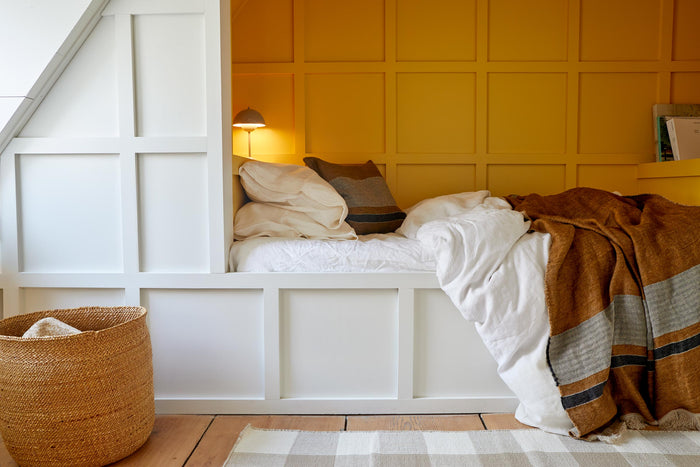 A cozy bed with white linens and pillows, complemented by a brown blanket is set against a paneled yellow wall. A lamp, basket, and white-painted wooden framing complete the space.