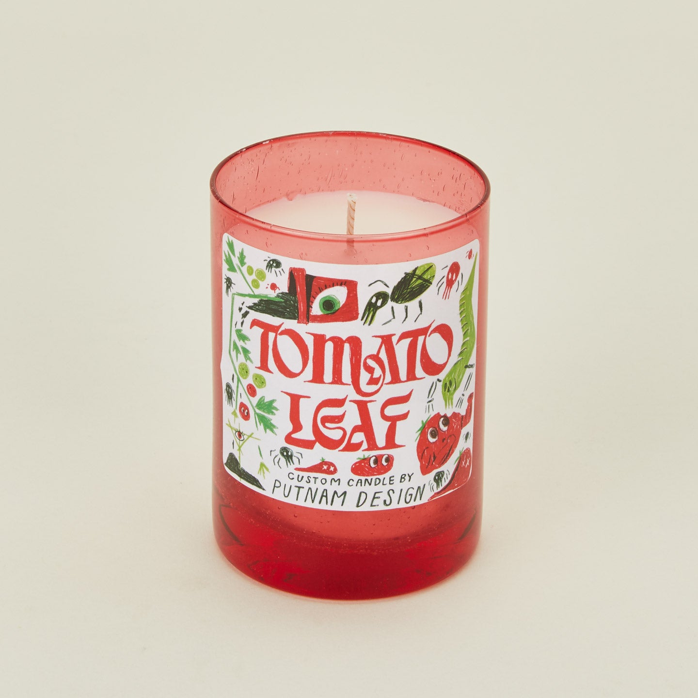 Candle with a red glass container and 'Tomato Leaf' label on a beige background