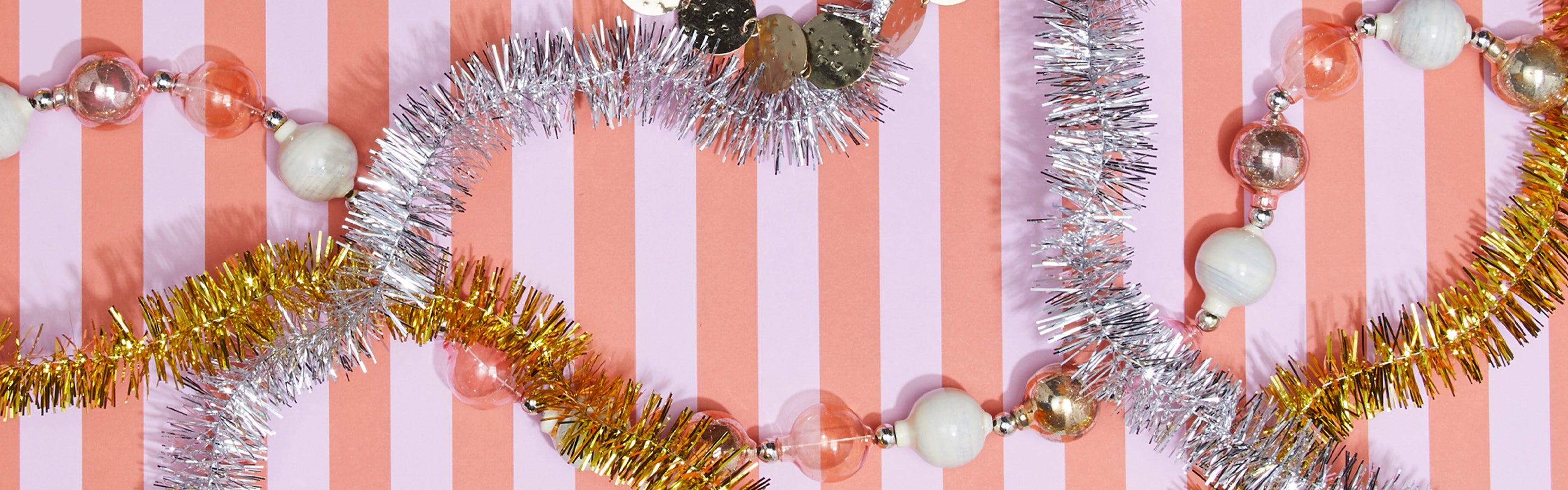 Shiny tinsel garlands with metallic and pearl-like beads are arranged on a pink and white striped background.