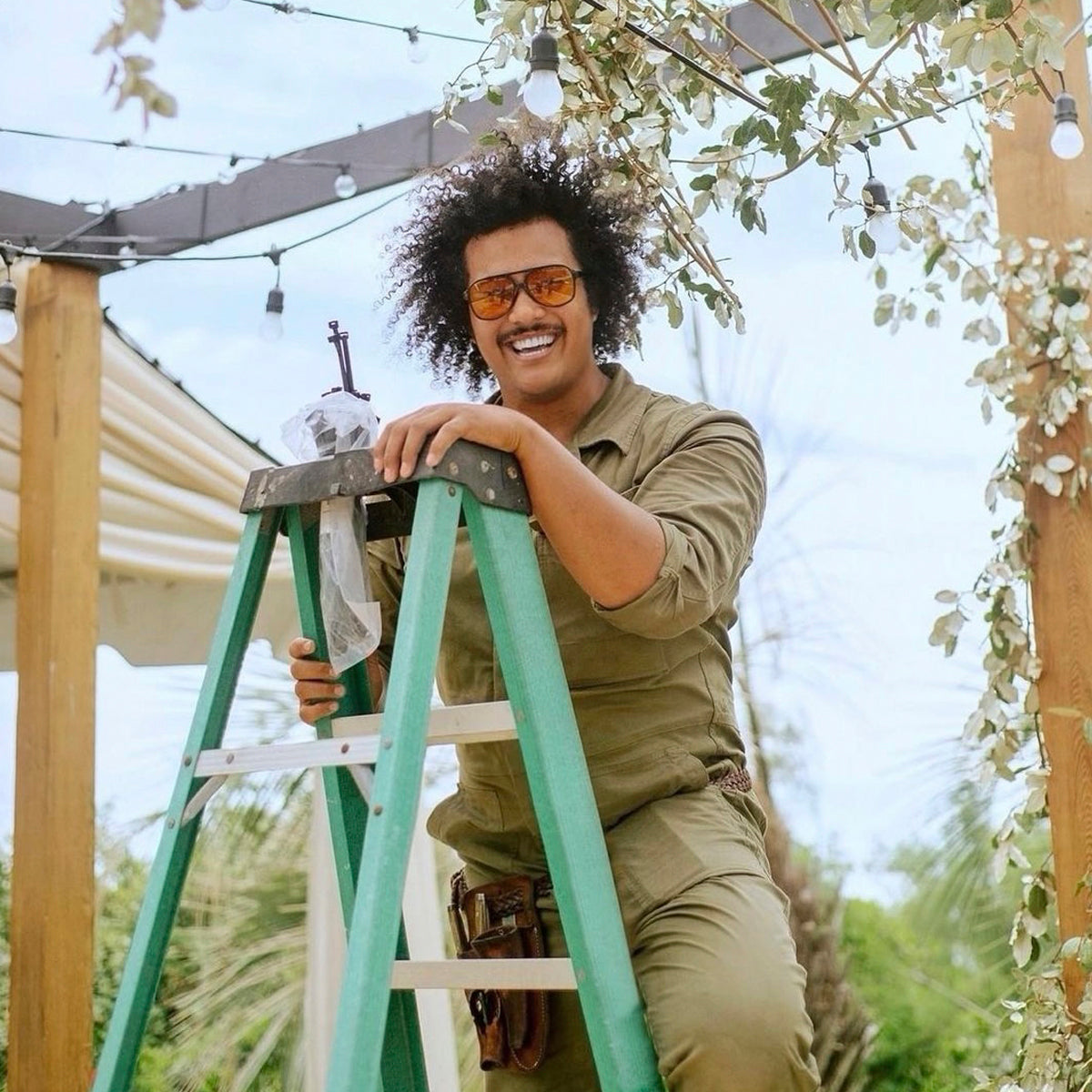 A person wearing sunglasses and a green jumpsuit stands on a ladder, smiling and working on overhead lights and greenery in an outdoor setting.