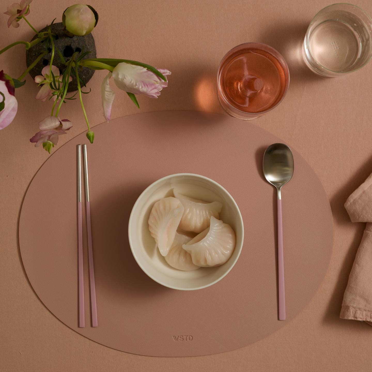 Dinner setting with a bowl of dumplings, pink chopsticks, spoon, and glasses on a pink placemat.
