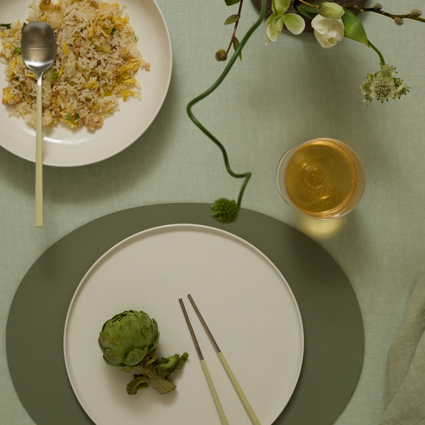 Table setting with a plate of artichoke, a glass of white wine, and a bowl of rice with a spoon.