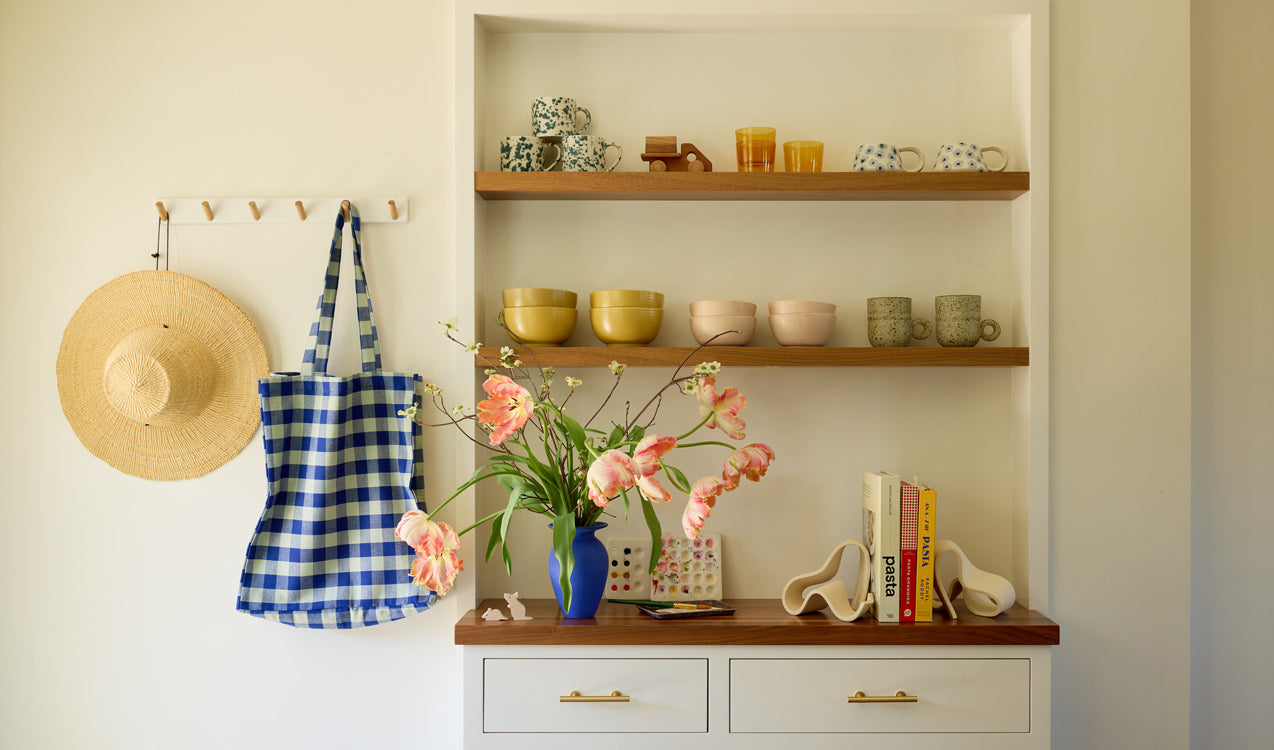 A built-in hutch with wood shelves above arranged with bowls and mugs in various colors. On the bottom shelf a blue vase full of pink flowers and a grouping of books held by sculptural ceramic bookends. Nearby a wall hook holds a woven straw hat and a blue gingham tote.